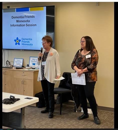 Two people standing at the front of a room. Screen with Dementia Friends information visible.